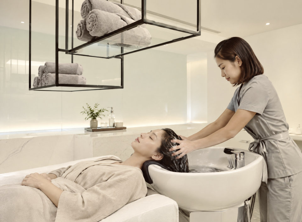 A massage professional performing a water-based scalp treatment beside a reclining guest at The Moment in Athens